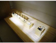 Sala conferenze/auditorium con palco in legno chiaro, lungo tavolo per relatori con sedie, microfoni e monitor, illuminato da un fascio di luce. A lato del palco c’è un piccolo podio con microfono, con pareti spoglie e un accesso a gradini.