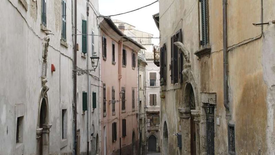Stradina stretta in un centro storico, pavimentata in sampietrini e fiancheggiata da alti edifici intonacati con persiane verdi e lampioni a parete. Lungo le facciate si vedono portali ad arco in pietra, muri leggermente usurati e cavi sospesi tra le case.