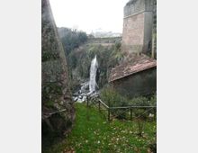 Veduta di una gola rocciosa con una cascata che scende tra pareti di pietra, vista da un sentiero erboso con staccionata in legno. Ai lati si notano murature in pietra e un edificio con tetto in tegole, con vegetazione sulle scarpate e costruzioni in lontananza.