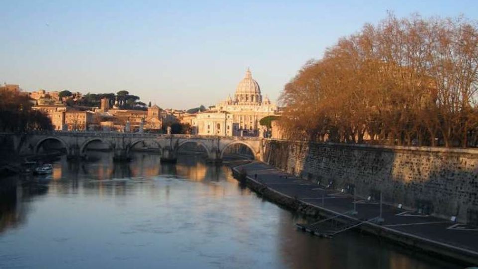 Veduta di un fiume urbano con un ponte in pietra ad archi e argini murati; sulla riva destra si vede un viale alberato spoglio. Sullo sfondo, tra gli edifici della città, spicca la grande cupola di una basilica sotto un cielo sereno.