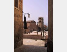 Veduta di una piazza in un borgo storico con pavimentazione in pietra, lampioni in ferro battuto e muretti, dominata da una chiesa in mattoni con campanile a torre e tetti in coppi. La scena è incorniciata da edifici alti ai lati e da alcuni cipressi lungo il percorso in discesa.