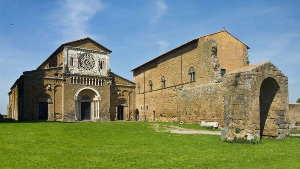 Veduta esterna di una chiesa in pietra in stile romanico con grande rosone e portale ad arco, affiancata da lunghi edifici monastici. In primo piano un prato verde e, sulla destra, un arco in muratura con resti di strutture in pietra sotto cielo sereno.