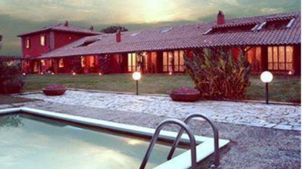 Piscina all’aperto con bordo in cemento e scaletta metallica in primo piano, affacciata su un vialetto ghiaioso e un prato con lampioni sferici. Sullo sfondo si vede un edificio basso con tetto in tegole e grandi vetrate illuminate, in un contesto rurale al tramonto.