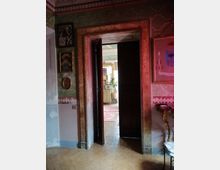 Interno di una casa storica con pareti decorate e cornice di porta in legno dipinto; una doppia porta socchiusa conduce a una stanza luminosa con tappeti e mobili. Sulle pareti sono appesi quadri e fotografie, e a destra si vede una piccola consolle con dettagli ornamentali.