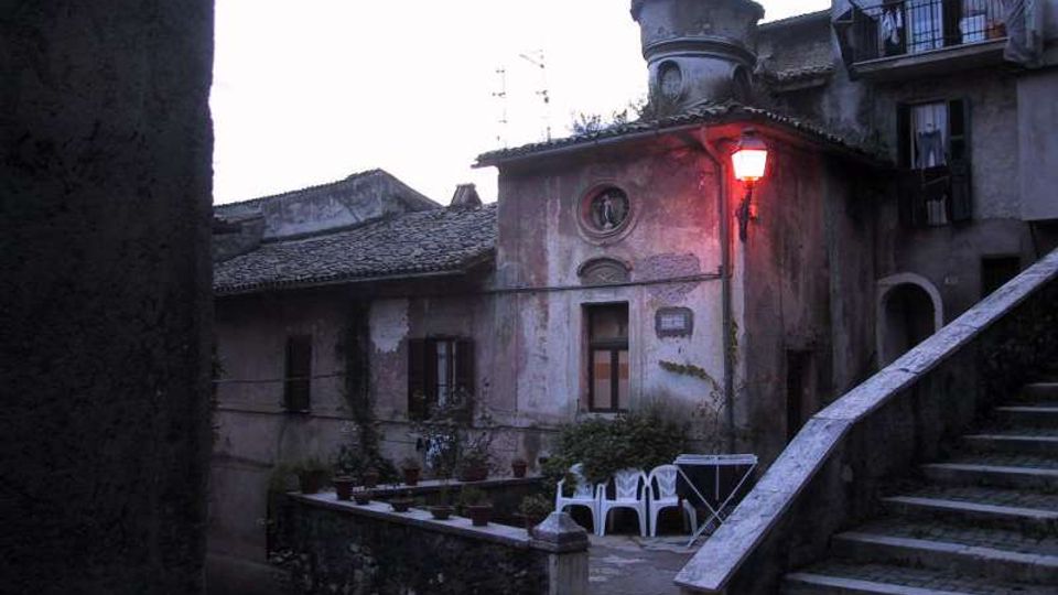 Cortile in un borgo storico con edifici in pietra intonacata e tetti in tegole, illuminato da un lampione rosso vicino a una piccola facciata con finestre tonde. In primo piano si vedono una scalinata in pietra con parapetto e alcune sedie e un tavolo in plastica su una terrazza.