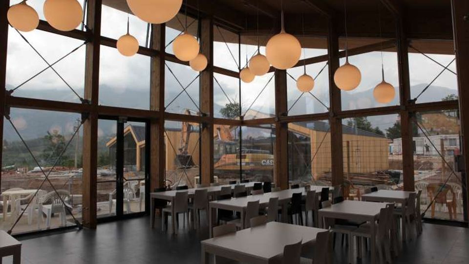 Interno di una sala ristorante moderna con grandi vetrate a tutta altezza incorniciate da travi in legno e tiranti metallici, con lampade sferiche sospese dal soffitto. Tavoli e sedie disposti in file su pavimento scuro, con vista verso un’area esterna e montagne sullo sfondo.