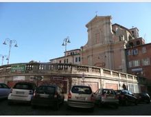 Vista urbana con un ponte in muratura con balaustra in pietra e lampioni, davanti a un parcheggio su strada. Sullo sfondo si vede una chiesa in stile barocco con facciata alta e edifici residenziali adiacenti sotto un cielo sereno.
