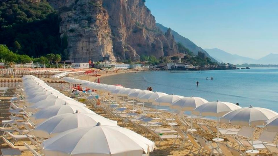 Spiaggia attrezzata con file di ombrelloni e lettini bianchi su sabbia dorata, delimitata da una staccionata bianca. Sullo sfondo si vede una grande falesia rocciosa che domina la costa e un mare calmo lungo la baia.