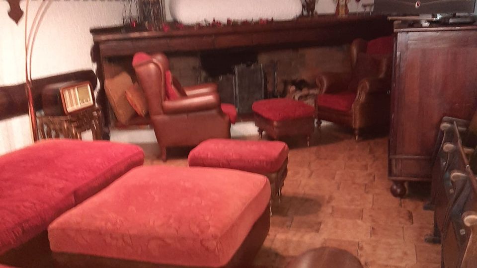 Salotto rustico con pavimento in piastrelle, grande camino in muratura con trave in legno e decorazioni, e lampadario sospeso in stile marocchino. Divani e poltrone in pelle con cuscini rossi e pouf sono disposti attorno al camino, con un mobile in legno e un televisore a lato.