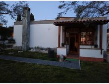 Facciata di una casa a un piano con pareti bianche e tetto in coppi, con portico d’ingresso sorretto da colonne in legno e grandi finestre con inferriate. Davanti c’è un piccolo giardino con prato, aiuole di pietre e un vialetto pavimentato che conduce alla porta, con un alto camino rivestito in pietra sul lato sinistro.