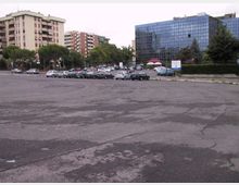 Ampio parcheggio asfaltato con numerose auto in sosta, circondato da alberi e aiuole. Sullo sfondo si vedono palazzi residenziali e un grande edificio moderno con facciata in vetro riflettente.