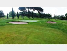 Green di un campo da golf su terreno ondulato, con due bunker di sabbia ai lati e una bandiera al centro. Sullo sfondo si vedono alberi alti e siepi che delimitano l’area.