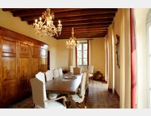 Sala da pranzo in stile classico con lungo tavolo in legno e sedie imbottite, soffitto con travi a vista e lampadari di cristallo. Grandi finestre con tende e pannellature in legno lungo una parete, con pavimento in piastrelle e vista verso una finestra sul fondo.