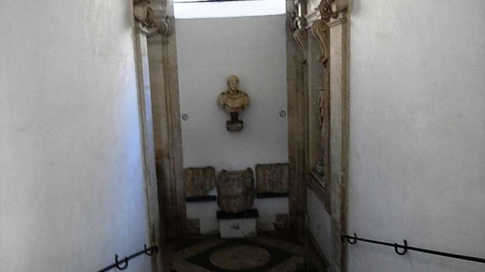 Scala interna in pietra che scende verso un piccolo pianerottolo, con pareti bianche e corrimano in ferro ai lati. In fondo si vede un arco con finestra semicircolare e decorazioni architettoniche in pietra, con un busto su mensola sulla parete.