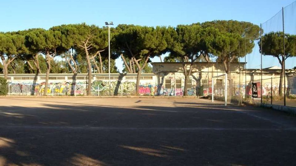 Campo sportivo all’aperto con ampia superficie asfaltata, recinzione metallica e rete alta su un lato. Sullo sfondo si vedono edifici bassi con muri coperti di graffiti, un lampione e una fila di pini sotto un cielo sereno.
