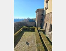 Giardino formale con siepi potate a labirinto all’interno di mura in pietra, accanto a un edificio storico con torre e facciata in muratura. Sullo sfondo si vedono colline alberate sotto un cielo sereno.