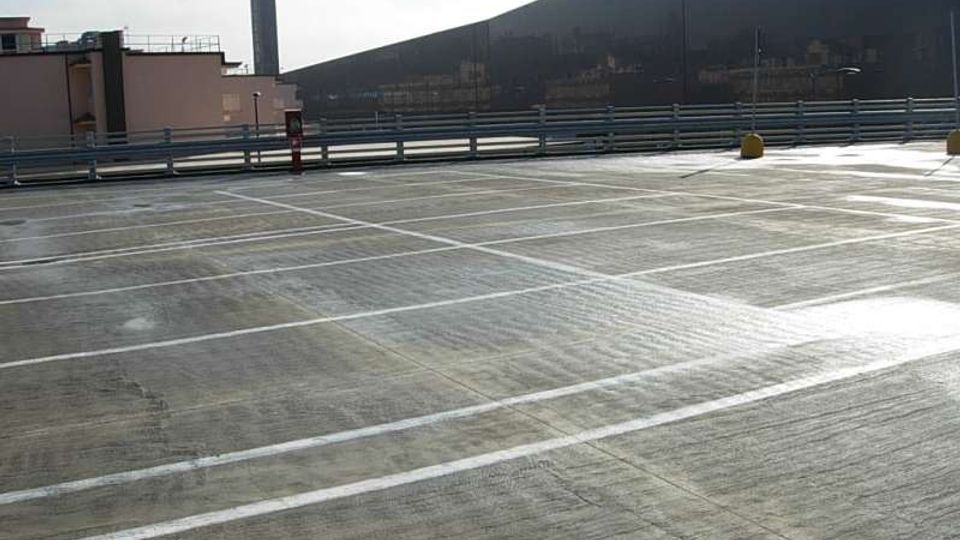 Parcheggio multipiano all’aperto con pavimentazione in cemento e strisce bianche che delimitano gli stalli, circondato da ringhiere metalliche. Sullo sfondo si vedono edifici industriali e una torre verticale sotto un cielo chiaro.