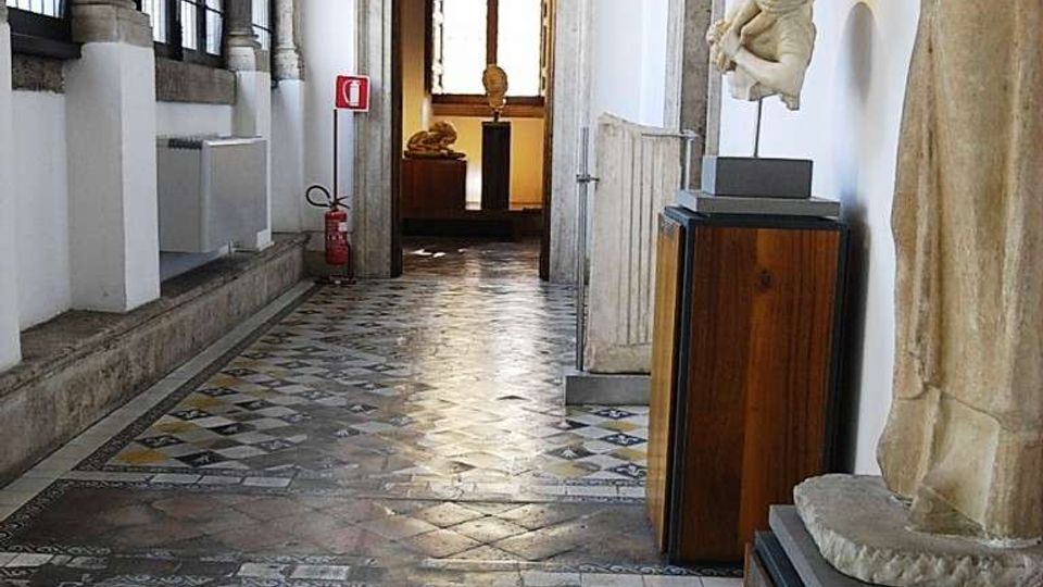 Corridoio interno di museo o palazzo storico con grandi finestre a griglia sul lato sinistro e pavimento a mosaico geometrico multicolore. Sulla destra sono presenti piedistalli con sculture e un passaggio con cornice in pietra che conduce a una sala sullo sfondo.