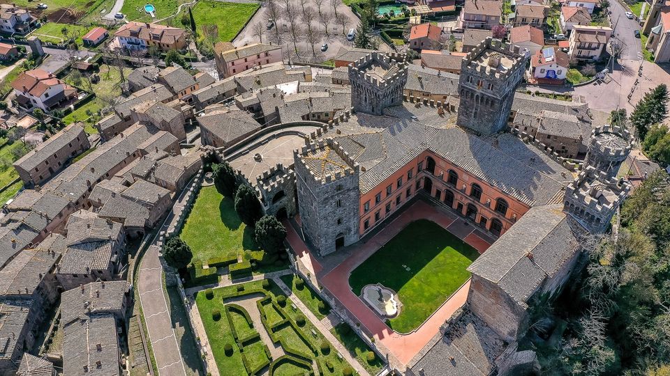 Veduta aerea di un borgo storico con un grande castello in pietra dotato di torri merlate, mura e un ampio cortile interno con prato e fontana. Intorno si vedono case con tetti in coppi e una strada che costeggia i giardini geometrici all’interno delle fortificazioni.