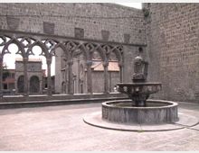 Cortile in pietra con una grande fontana circolare al centro, circondato da alte mura in blocchi di tufo. Sul lato sinistro si vede una loggia con archi gotici e colonnine che incorniciano edifici storici sullo sfondo.