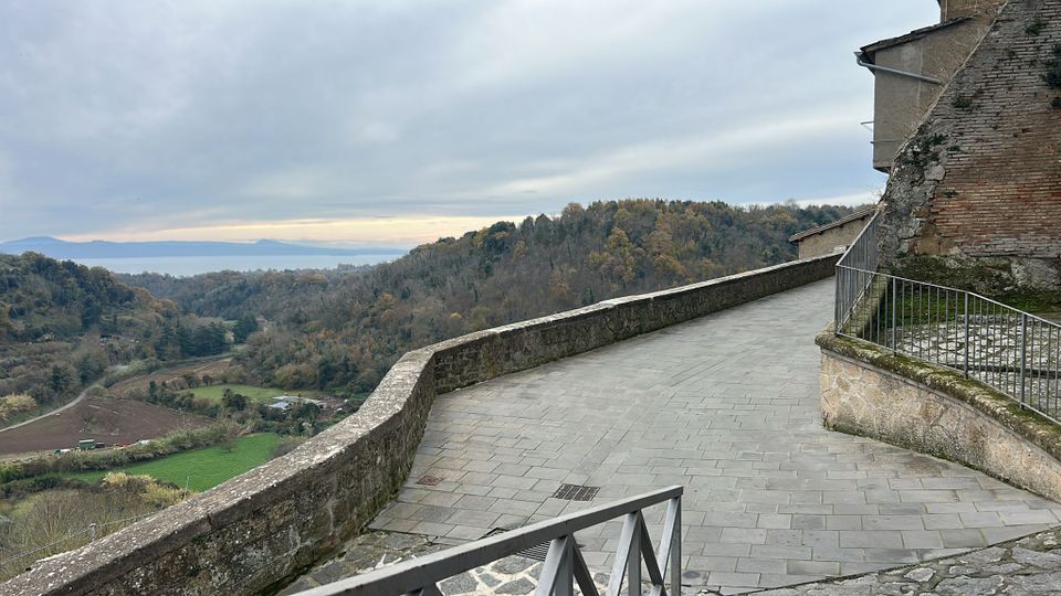 Terrazza panoramica lastricata in pietra con muretto di contenimento, affiancata da un edificio in muratura e una ringhiera metallica. Sullo sfondo si apre una vista su colline boscose, campi nella valle e un lago in lontananza sotto un cielo nuvoloso.