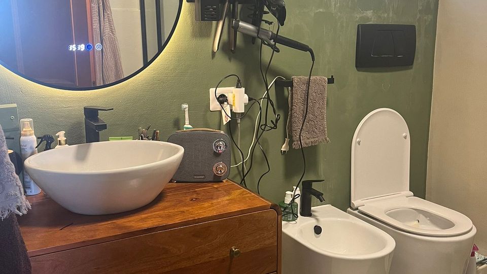 Bagno moderno con pareti verde salvia, mobile lavabo in legno con lavandino a ciotola e grande specchio rotondo retroilluminato. A destra si vedono bidet e WC sospesi sotto una finestra satinata, con accessori a parete e tappetino sul pavimento chiaro.