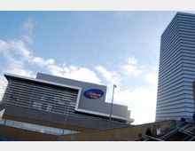 Edificio moderno di un complesso commerciale con facciata grigia a pannelli e ampie vetrate, con un grande logo ovale “UCI Cinecittà” in evidenza. A destra si vede una torre alta rivestita da fasce orizzontali e, in primo piano, una scalinata esterna con parapetti e lampioni sotto un cielo parzialmente nuvoloso.