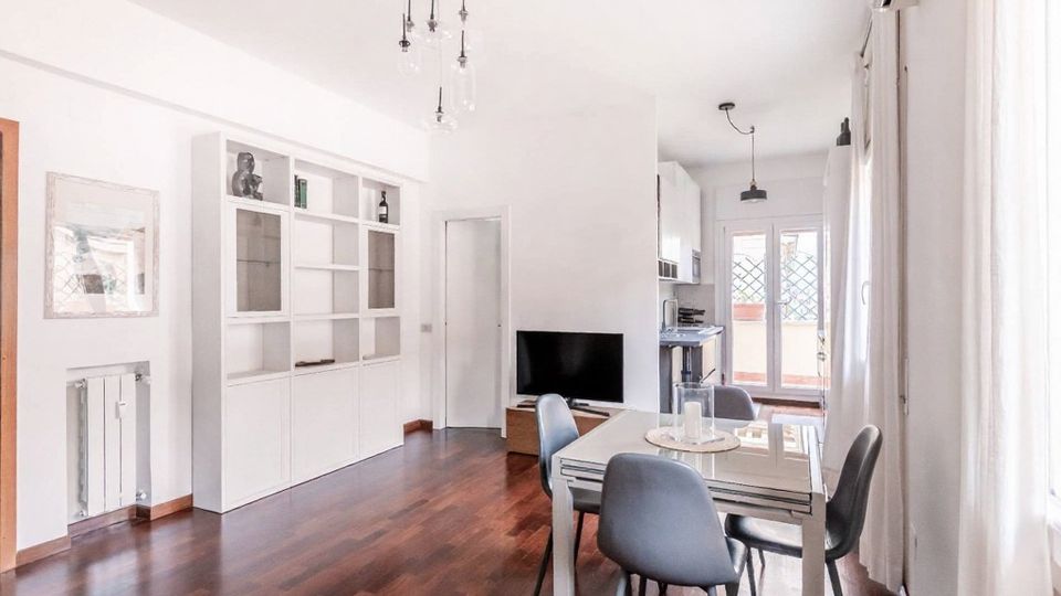 Soggiorno open space luminoso con pavimento in parquet scuro, tavolo da pranzo con sedie grigie, libreria a parete bianca e TV vicino al passaggio verso la cucina. Ampie finestre con tende chiare e portafinestra sul fondo che conduce a un balcone.