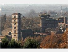 Complesso storico in pietra con una torre campanaria quadrata a più livelli e edifici annessi dai tetti in coppi, circondato da alberi autunnali. Sullo sfondo si vede una campagna pianeggiante leggermente nebbiosa con campi e strutture sparse.