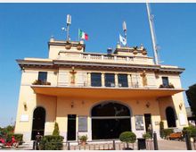 Edificio pubblico a due piani color crema con grande ingresso ad arco centrale, balconata con finestre e terrazza superiore con balaustra. Sul tetto sono presenti antenne e aste con bandiere, con piccolo spazio esterno recintato e panchine davanti alla facciata.