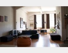 Ampio soggiorno in stile loft con pavimento in legno, divano scuro e pouf, pareti con quadri e lampade a sospensione. Sul fondo grandi finestre a riquadri con tende scure illuminano la stanza, con una piccola area studio e una pianta vicino alla finestra.