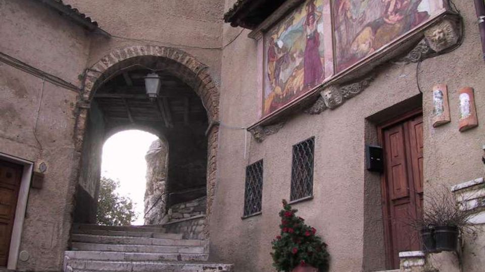 Scalinata in pietra che conduce a un arco in muratura con lanternino, tra edifici intonacati. Sulla facciata a destra sono visibili grandi affreschi sotto il tetto, finestre con inferriate e una porta in legno.