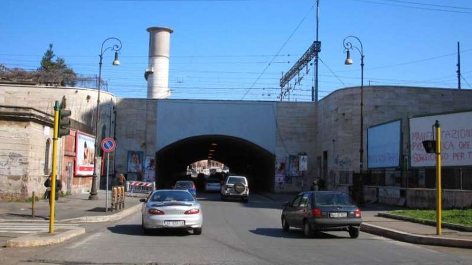 Strada urbana che conduce a un ampio sottopasso in muratura con grande arco centrale, pareti con cartelloni e una ciminiera sullo sfondo. Sopra il tunnel si vedono cavi della linea ferroviaria e lampioni stradali ai lati della carreggiata.
