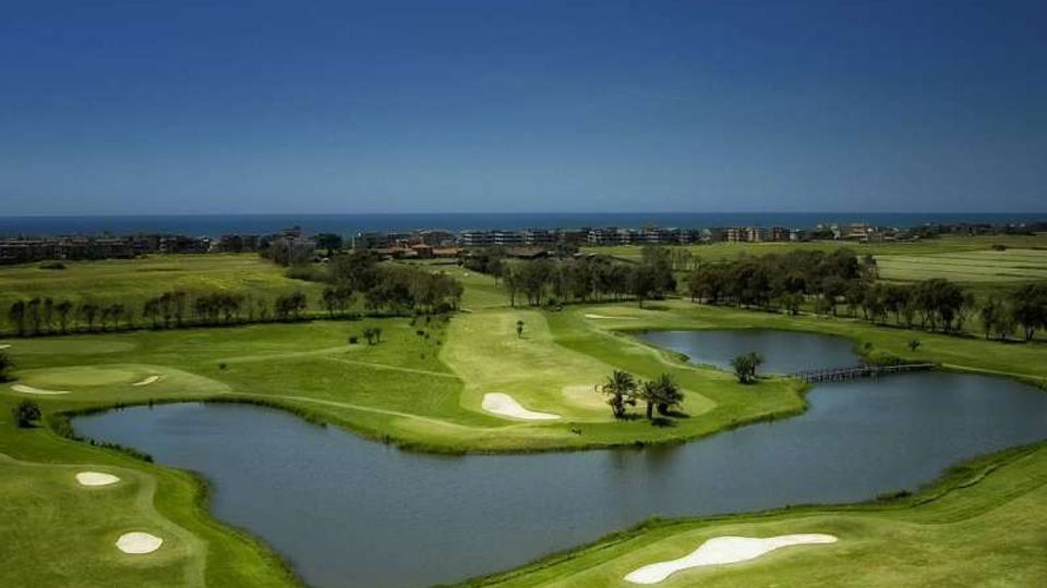 Campo da golf con ampi fairway verdi, bunker di sabbia e due laghi che circondano un green collegato da un piccolo ponte. Sullo sfondo si vede la costa con il mare all’orizzonte e alcuni edifici bassi sotto un cielo limpido.