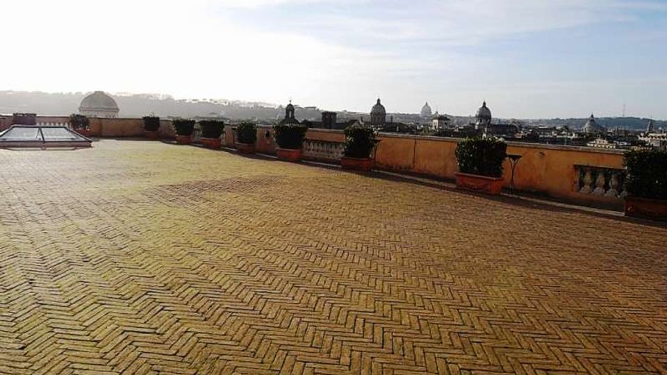 Ampia terrazza panoramica con pavimentazione in mattoni a spina di pesce e parapetto in muratura, fiancheggiata da vasi con siepi. Sullo sfondo si vede lo skyline urbano con cupole e campanili sotto un cielo chiaro.