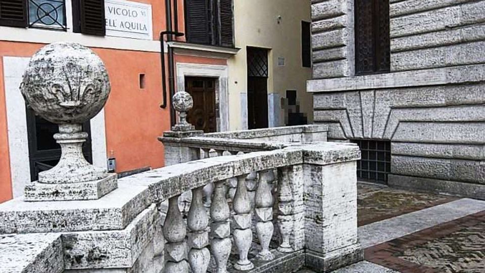 Vicolo urbano tra edifici storici con facciate color ocra e arancione e un palazzo in pietra bugnata con finestra ad arco. In primo piano un parapetto in travertino con balaustre e sfere decorative, accanto a una pavimentazione in pietra e mattoni con motivo circolare.