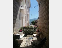 Piccola terrazza con sedute in rattan e tavolino su pavimento a motivi geometrici, delimitata da una ringhiera con fiori. L’area è incorniciata da un grande arco tra due pareti decorate e si affaccia su una strada del borgo con montagne sullo sfondo.