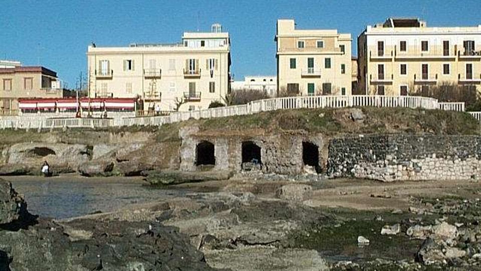 Veduta di un lungomare con edifici chiari a più piani affacciati sulla costa, separati da una staccionata bianca. In primo piano si vedono scogli e piattaforme rocciose con piccole grotte alla base di un muretto in pietra.