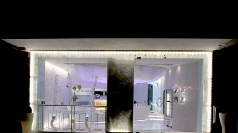 Padiglione moderno con facciata vetrata e cornice luminosa, che mostra un interno bianco e minimalista con bancone e arredi essenziali. All’esterno si vede una breve fascia di prato davanti all’ingresso.