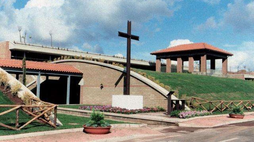 Area esterna di un complesso commemorativo con una grande croce in legno su basamento, vialetti pavimentati e aiuole fiorite. Sullo sfondo si vedono edifici in mattoni con tetti in tegole e un pendio erboso con staccionate in legno.