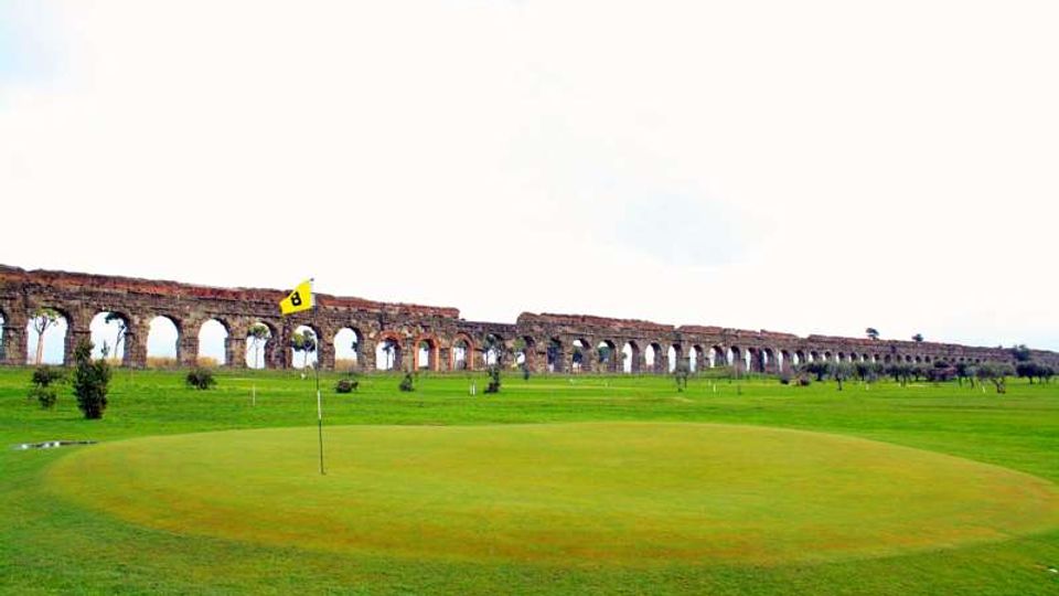 Campo da golf con green e bandierina in primo piano, circondato da ampie distese erbose. Sullo sfondo si estende un lungo acquedotto in mattoni con una serie di arcate, sotto un cielo chiaro.