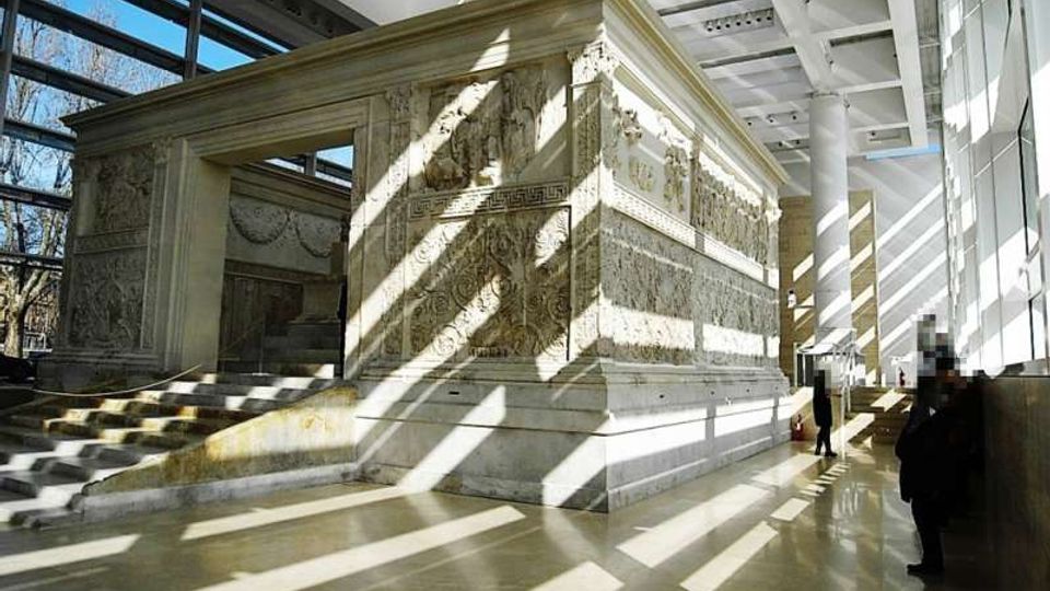 Interno di un museo moderno con ampie vetrate e soffitto a griglia, che ospita una grande struttura in pietra scolpita con bassorilievi e una scalinata d’accesso. La luce naturale crea lunghe ombre geometriche sul pavimento lucido.