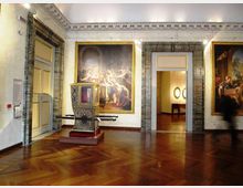 Sala museale con pavimento in parquet lucido, grandi dipinti incorniciati alle pareti e una portantina storica decorata esposta al centro. Porte e passaggi con cornici in marmo conducono a un’altra stanza illuminata.