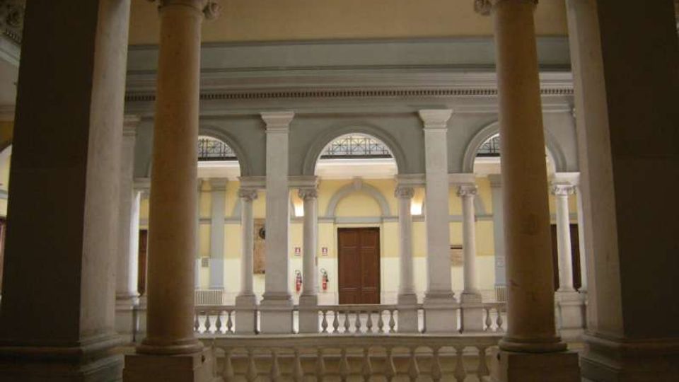 Cortile interno di un edificio storico con colonne in pietra e ballatoio con balaustra, affacciato su una facciata con arcate e finestre ad arco. Al centro si vede una porta in legno e pareti dai toni chiari sotto un soffitto alto.
