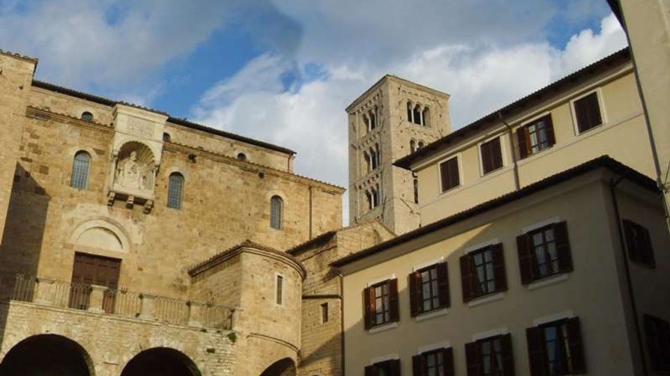 Veduta di una piazza con una grande chiesa romanica in pietra, con portale ad arco, loggia e arcate alla base, affiancata da un campanile quadrato. Sul lato destro si vede un edificio residenziale chiaro a più piani con persiane scure, sotto un cielo parzialmente nuvoloso.