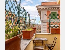 Terrazza urbana con muretto perimetrale e grigliati verdi, fioriere rettangolari in terracotta e un piccolo tavolo con sedia pieghevoli in legno. Sullo sfondo si vedono facciate di edifici con persiane verdi e cielo parzialmente nuvoloso.