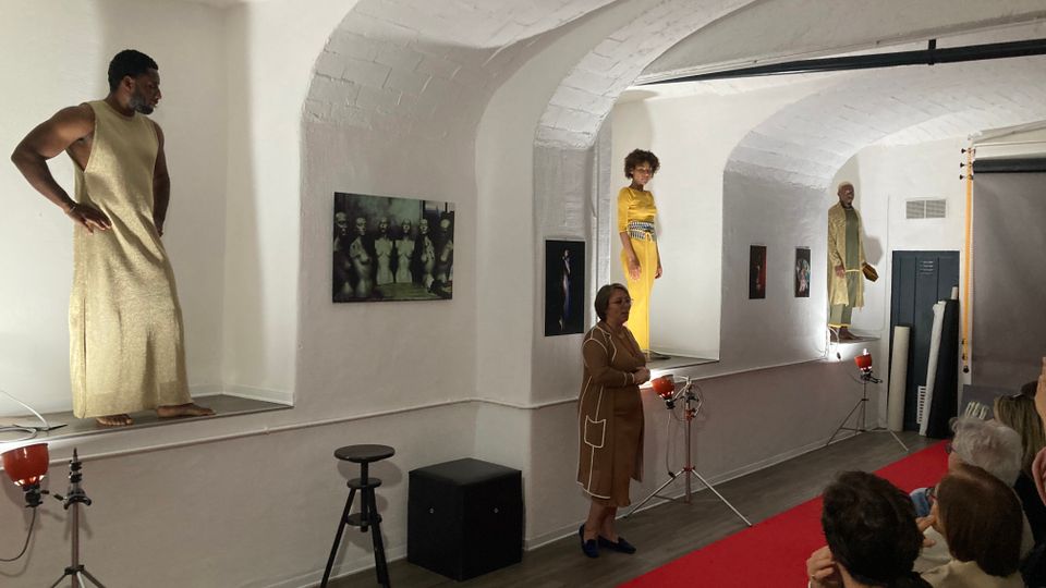 Interno di una galleria con pareti bianche e soffitto a volte in mattoni, con una lunga mensola rialzata che ospita fotografie appese e manichini in abiti da passerella. Sul pavimento in legno corre un tappeto rosso, con luci su treppiede e attrezzature fotografiche lungo il lato.