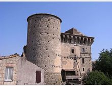 Antico edificio fortificato in pietra con grande torre cilindrica e struttura adiacente con loggiato ad archi, affacciato su un piccolo nucleo di case. Sullo sfondo si vede il cielo sereno e vegetazione ai margini dell’abitato.