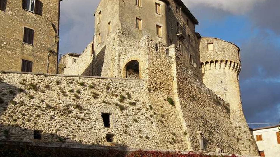 Castello medievale in pietra con alte mura merlate e una torre cilindrica, affiancato da edifici in muratura con finestre e tetti spioventi. In primo piano si vede un pendio con vegetazione e una fascia di rampicanti rossi alla base delle mura, sotto un cielo parzialmente nuvoloso.