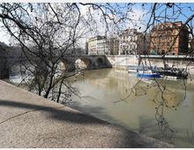 Veduta di un fiume cittadino con un ponte in pietra a più arcate, edifici storici lungo la sponda e alcune barche ormeggiate. In primo piano si vedono rami spogli di alberi e un parapetto in pietra del lungofiume.